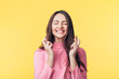 Young beautiful woman crossing her fingers and wishing for good luck over yellow background