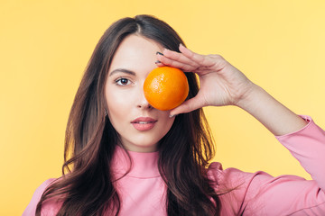 Wall Mural - Beautiful young woman holding orange in front of eye isolated on yellow background