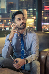 Wall Mural - Handsome young businessman with earphones listening to music on smart phone while sitting in comfortable chair with city skyline buildings in the background