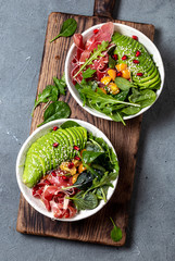 Wall Mural - Low carbs bowl. Fresh salad with green spinach, rucola, avocado an ham serrano in white bowl, gray background, top view