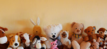 Soft Plush Toys For Children Sitting In Row, Different Toy Animals For Kids On Yellow Wall Background With Copy Space As Horizontal Banner. 