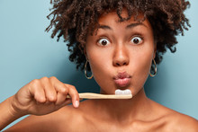 Cropped Shot Of Surprised Dark Skinned Woman With Curly Hair, Keeps Toothbrush Near Folded Lips, Whitens Teeth, Cares Of Oral Hygiene, Has Healthy Skin, Isolated Over Blue Background. Dental Care