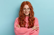 Satisfied ginger charming woman with curly hair, wears transparent glasses, keeps arms folded over chest, has pleasant talk with contemporary, wears oversized clothes, isolated over blue background