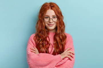 Satisfied ginger charming woman with curly hair, wears transparent glasses, keeps arms folded over chest, has pleasant talk with contemporary, wears oversized clothes, isolated over blue background