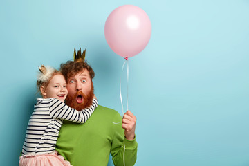 Happy Fathers Day! Positive child and dad have funny time together at home, play with air balloon, wear crowns, pose over blue studio wall with empty space for your advert. Family celebration