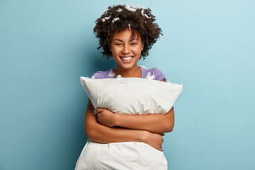 Photo of happy dark skinned woman cuddles white pillow, smiles broadly after awakening in morning, glad to have pillow fight with husband, has feathers in curly hair, poses over blue background