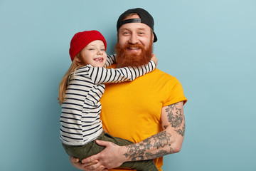 Wall Mural - Isolated shot of happy father with ginger beard, yellow t shirt, carries small daughter, recieves hug with love, spend free time together in family circle, express positive emotions. Parents, children