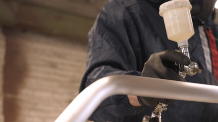 In slow motion a professional man (guy) in a garage (service) in working clothes dressed in a sweater sets up a paint gun (spray gun) and starts painting (putting a layer of ceramics) a yacht.