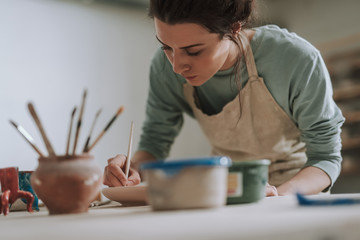 Skillful young woman in apron painting pottery at workshop