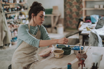 Serious young woman in apron painting pottery at workshop