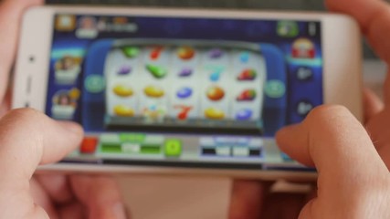 Casino slot machine app on mobile screen against  hand of man using smartphone. Gambling addicted man in front of online casino slot-machine on mobile phone. Screen is blurred. Closeup. 4K UHD.