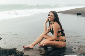 Side view of sexy girl in bikini spends time on black sand beach. Young woman with long hair posing outdoor