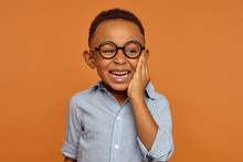 Emotional Black Dark-skinned Little Boy Grimacing And Holding Hand On His Cheek Because Of Intolerable Toothache. Handsome Cheerful Dark Skinned African Child In Glasses And Shirt Smiling Broadly