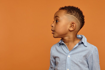Wall Mural - Waist up up image of handsome adorable dark skinned little child with stylish hairdo having emotional facial expression, standing isolated at blank orange wall with copyspace for your advertisement