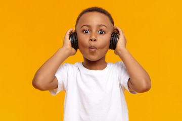 Wall Mural - Picture of stylish cool Afro American schoolboy making funny facial expression, being excited with amazing track while listening to music using wireless black headphones, posing isolated in studio