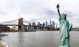 Fototapeta Nowy Jork - Panorama view of The Statue of Liberty with Brooklyn Bridge and Manhattan downtown sky scraper with cloud blue sky background, Financial district lower Manhattan, New York City, USA.
