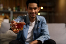 Close Up Shot Of Asian Man Wearing Jean Jacket Sitting On Sofa Holding Glass Of Whiskey In Night Club Restaurant. Focus On The Glass. Alcohol Drinking Concept.