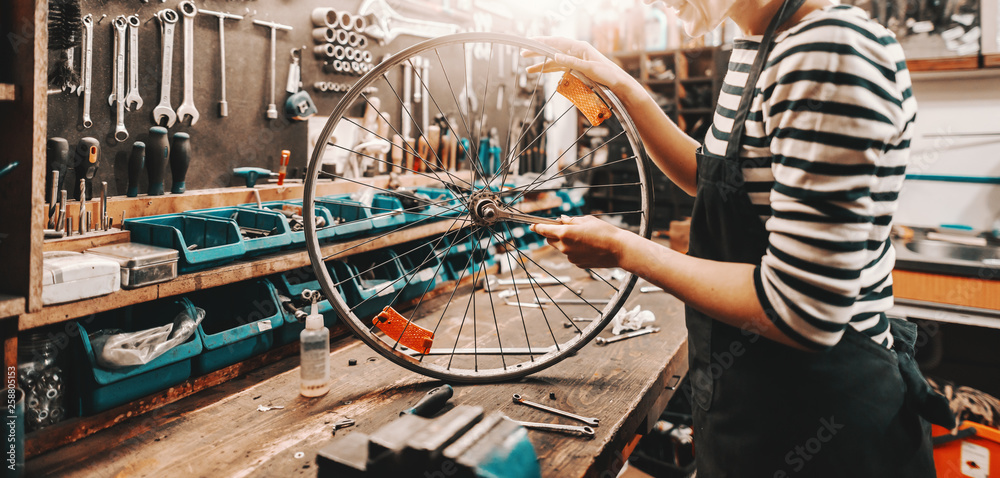 Cute Caucasian female worker holding and repairing bicycle wheel while standing in bicycle workshop. - obrazy, fototapety, plakaty 