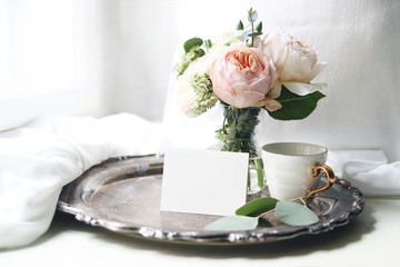 Wall Mural - Spring, summer still life. Blank place card mockup, cup of coffee on old silver tray near window. Vintage feminine styled photo. Floral composition of pink English roses, Ranunculus and eucalyptus.