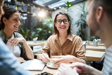 Group of positive creative young entrepreneurs in casual clothing sitting in circle at table in modern cafe and discussing project development