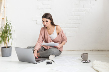 A young happy woman sits on the floor in a bright apartment or office interior and works at a laptop, freelancer girl at work, the concept of distance learning students, paperwork and online learning