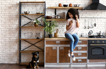 Wall Mural - African-American woman with cute funny dog drinking coffee in kitchen