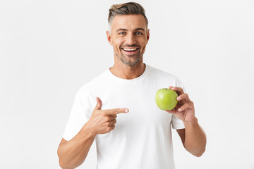 Wall Mural - Portrait of attractive man 30s having bristle in casual t-shirt posing on camera and holding green apple in hand