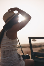 Young Woman On A Desert Safari Holding Her Hat And A Camera, Sitting On A Vintage Car With The Sun In The Background.
