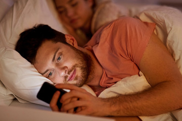 technology, internet addiction and cheat concept - man using smartphone at night while girlfriend is sleeping