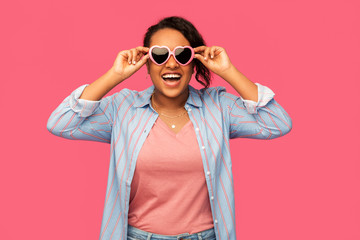 summer, valentine's day and eyewear concept - happy african american young woman in heart-shaped sunglasses over pink background