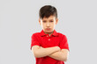 childhood, expressions and people concept - displeased little boy with crossed arms in red polo t-shirt pouting and squinting over grey background