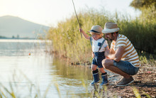 A Mature Father With A Small Toddler Son Outdoors Fishing By A Lake.