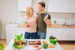 © estradaanton - Vegan happy loving couple holds bottles with natural smoothie while cooking raw vegetables in white kitchen. Diet detox. Happy family. Raw food diet. Vegetarian food.