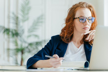 Serious Experienced Female Coach Makes Records In Notepad, Focused At Screen Of Laptop Computer, Has Long Red Hair, Wears Transparent Glasses, Watches Webinar Online. Business And Job Concept