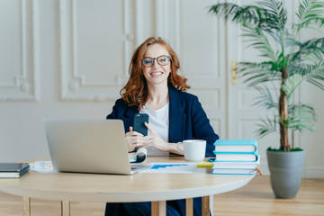 Wall Mural - Horizontal shot of optimistic woman installs application on modern cell phone, checks notification, surrounded with paper documents, works on laptop computer, has red hair, dressed formally.