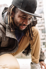 Wall Mural - Handsome African American with dreadlocks in brown trousers and a blue sweater in a black cap on his head against the background of houses, city