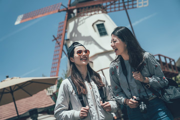 Wall Mural - two joyful asian women tourist travel with camera in solvang santa barbara california america. young girl best friends travel together smiling sightseeing windmill outdoor under blue sky in summer