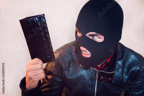 Fotomural Young ill-intended man wearing black mask holds the black leather women purse and looks to his loot