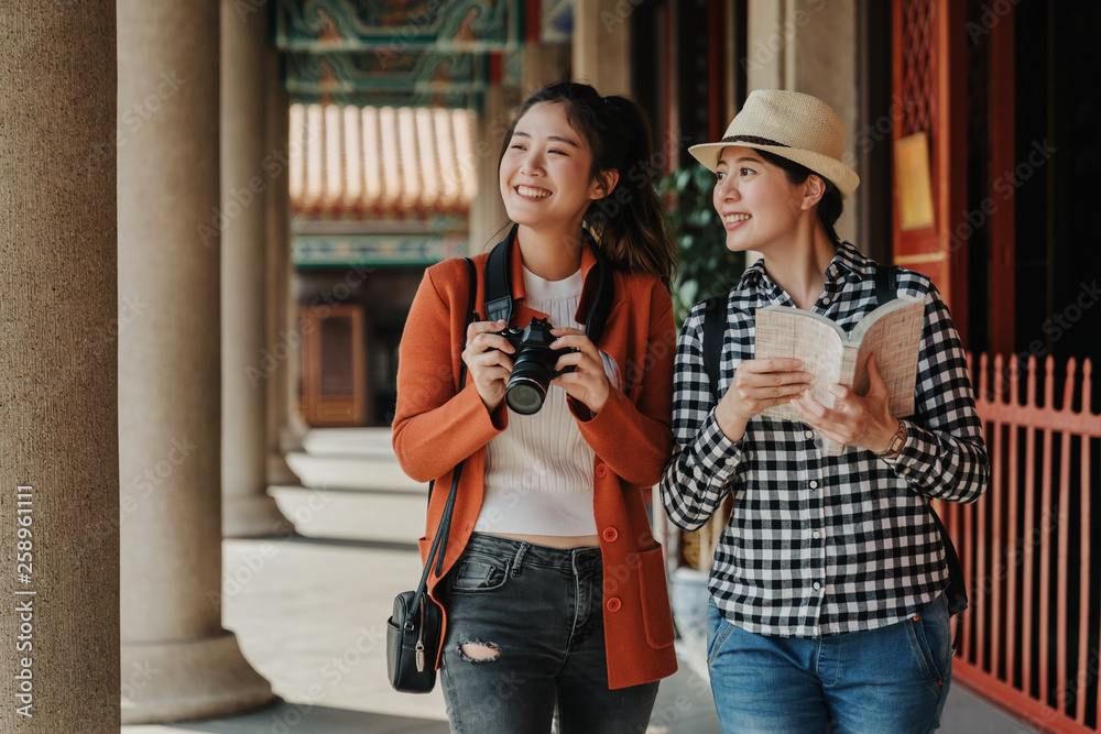 two girls tourist visiting bangkok maining attractions in thailand. women friends travel walking in corridor hallway in chinese temple on sunny day in spring. backpackers smiling with camera and book - obrazy, fototapety, plakaty 