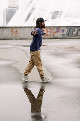 Wall Mural - Handsome African American with dreadlocks in brown trousers and a blue sweater in a black cap on his head against the background of houses, city