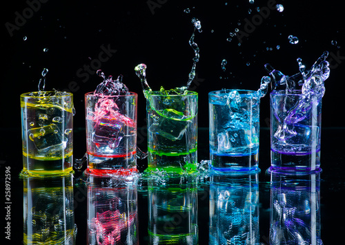 Fotomural  Multicolored glasses filled with alcoholic drinks, with splases of ice cubes falling inside, standing on the mirror surface