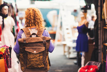 Blonde Curly Hair Woman Backpacker Traveler Viewed From Rear At Used Market Enjoying The Shopping And Alternative Vacation - Leather Backpack View For Travel Concept