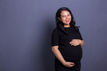 Portrait of happy attractive Asian mother with her pregnant belly smiling at camera, standing against grey background