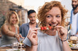 © Westend61 - Young woman eating meat skewer at a backyard party with friends