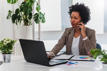 Wall Mural - Happy smiling african-american business woman working on laptop at office, using smart phone. Businesswoman sitting at her working place