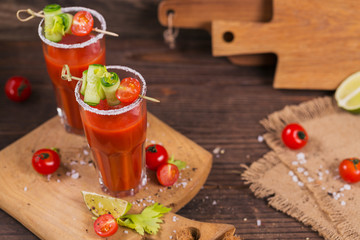 Two glasses of tomato juice decorated with fresh tomatoes, cucumber and leaves on a wooden background