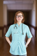 Smiling portrait of white redhead woman doctor with stethoscope on neck and hands in the pockets