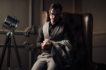 Wall Mural - Young handsome man posing for a fashion shoot in a studio. Fashion as a lifestyle. Man wearing a beard. Model sitting on a sofa in tuxedo. Successful fashionable businessman. Business look advertising