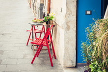 Red Table And Chair In An Outdoor Café With Blue Wooden Door In Syracuse (Siracusa) Sicily.