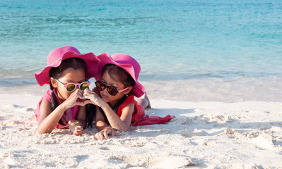 Wall Mural - Two cute asian child girls wearing pink hat and sunglasses playing with sand and making hand heart shape together on the beach near the beautiful sea in summer vacation